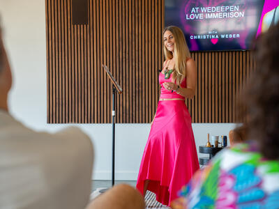 Christina Weber presenting at September 2025 WeDeepen Love Immersion in pink dress with branded screen